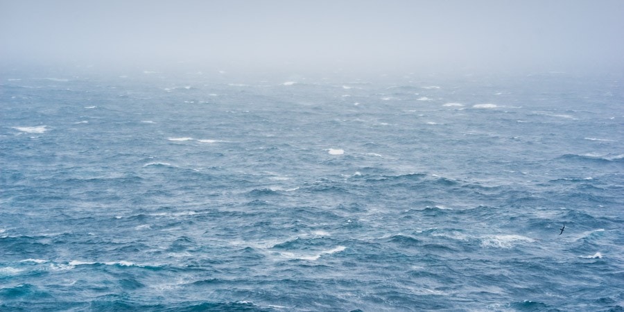 Choppy ocean waters of the Southern Ocean stretch to the horizon under a misty, overcast sky, with small whitecaps visible across the surface. A single seabird can be seen flying low over the waves, emphasizing the remote and stormy conditions at sea.