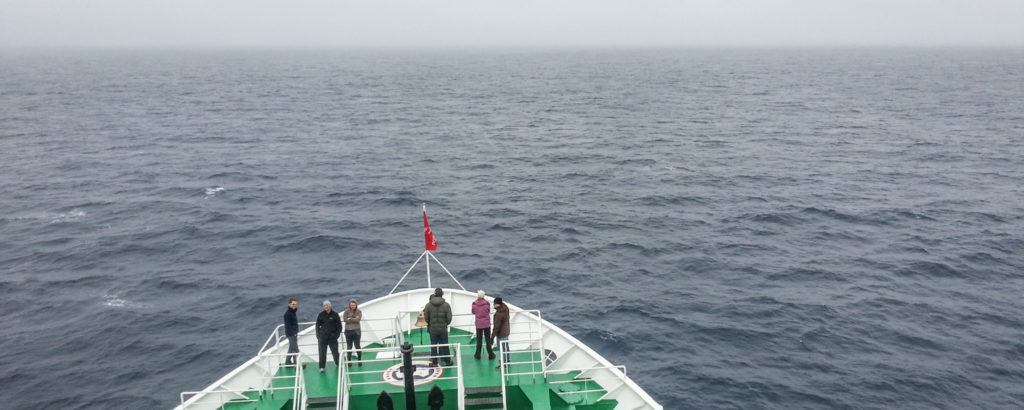Passengers stand at the bow of a ship cruising through choppy, gray waters under an overcast sky. The wide-open ocean view emphasizes isolation and movement, capturing the potential for motion sickness on an Antarctica cruise.