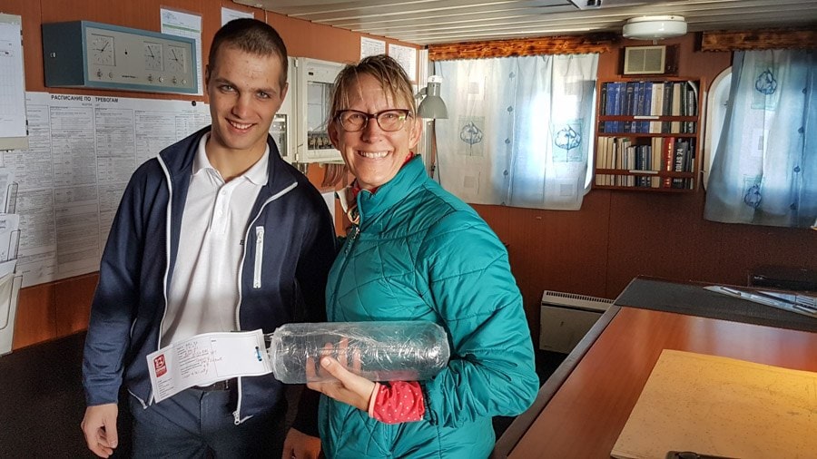 Two people stand inside a ship’s cabin smiling at the camera, with one of them holding a clear cylindrical sampling container labeled with a white and red tag. The setting features charts, bookshelves, and windows, indicating a workspace for navigation or research operations.