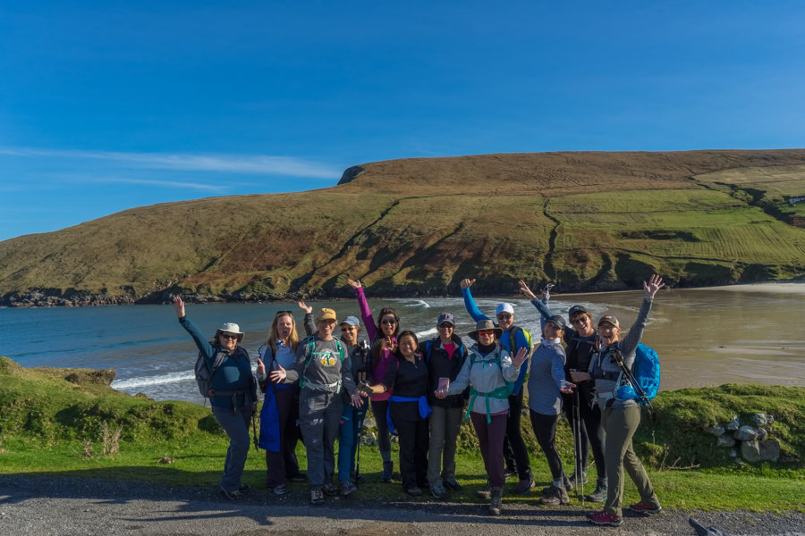 This image shows a group of hikers posing together in front of a scenic coastal landscape. The group, consisting mostly of women dressed in outdoor gear and backpacks, is standing on a grassy path with the ocean, sandy shoreline, and rolling green hills behind them. They have their arms raised in excitement, smiling under a clear blue sky, suggesting a joyful moment during an outdoor adventure. 