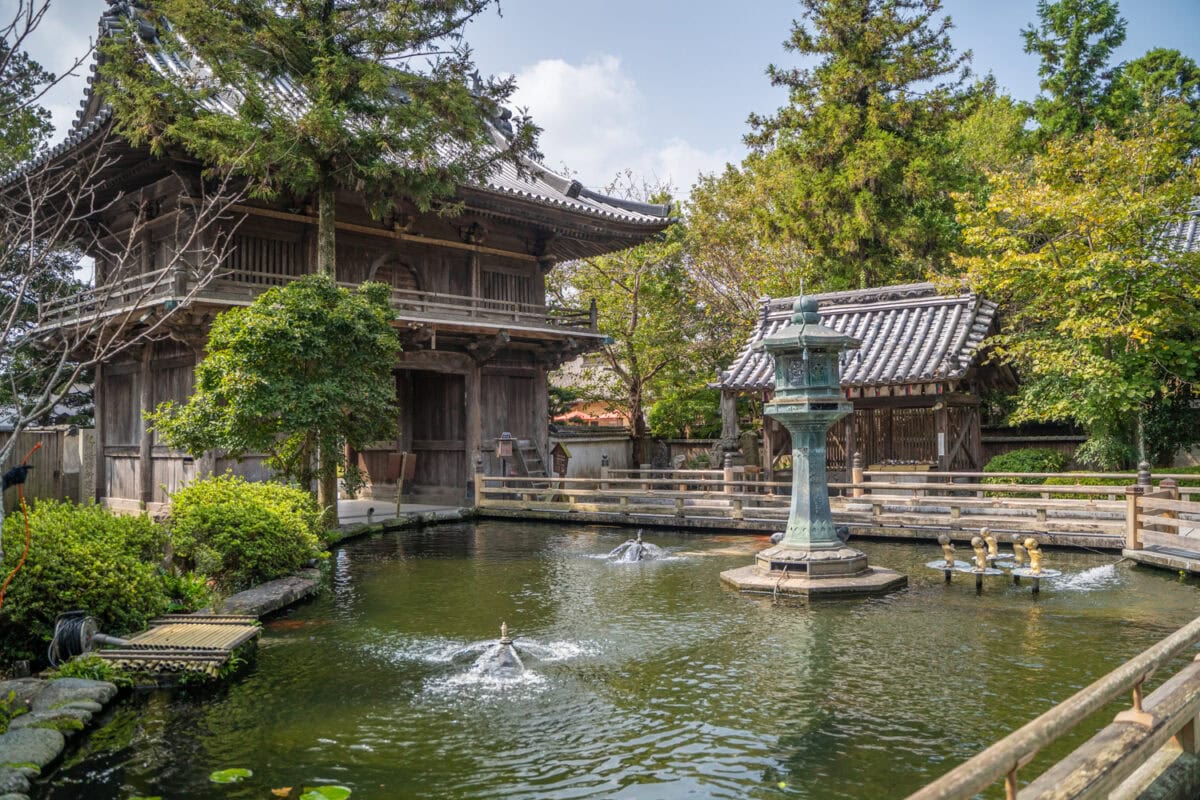 The image showcases Temple 1 Ryozen-ji, a tranquil Japanese temple garden featuring a serene pond with small water fountains and a traditional stone lantern in the center. Surrounding the pond, lush greenery and meticulously arranged plants complement the rustic wooden temple structures with sloped tiled roofs in the background. The scene exudes a peaceful atmosphere, highlighting the harmony between natural and architectural beauty in a traditional Japanese setting.
