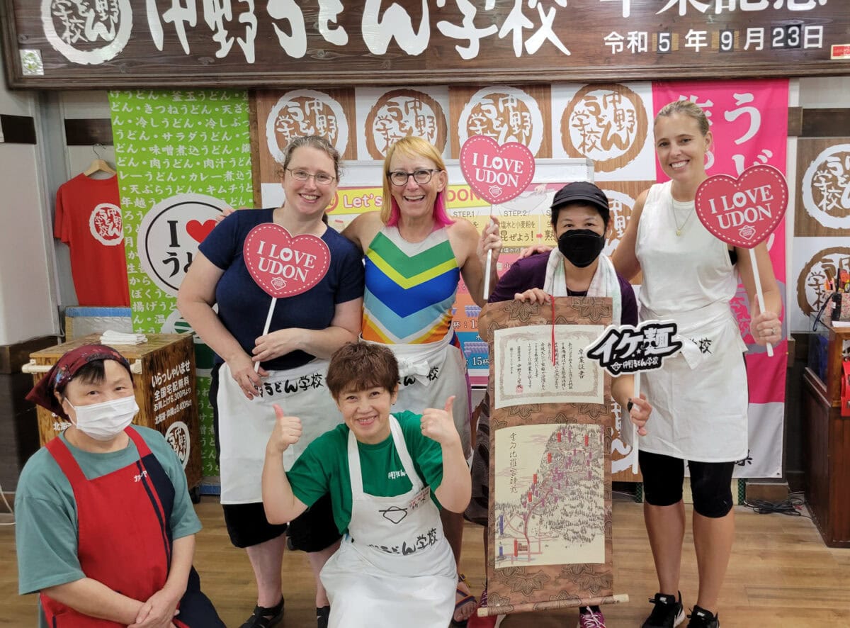 A group of smiling individuals, wearing aprons and holding "I Love Udon" signs, pose together at an udon cooking class. They stand in front of colorful banners and signs, including one person displaying a large scroll with Japanese text and map illustrations. The atmosphere is joyful, capturing the fun and pride in making udon noodles together.