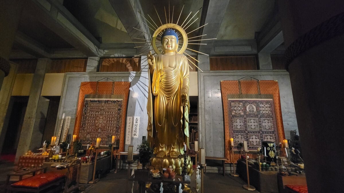 The image features Temple 6 Anrak on Shikoku Island, a grand golden statue of a Buddha, adorned with a radiant halo and standing in a serene mudra pose, inside an ornately decorated temple hall. Intricate tapestries hang on the walls, and offerings, candles, and decorative items are arranged at the statue's base, creating a reverent and sacred atmosphere. The lighting accentuates the Buddha's peaceful expression and the spiritual ambiance of this worship space.