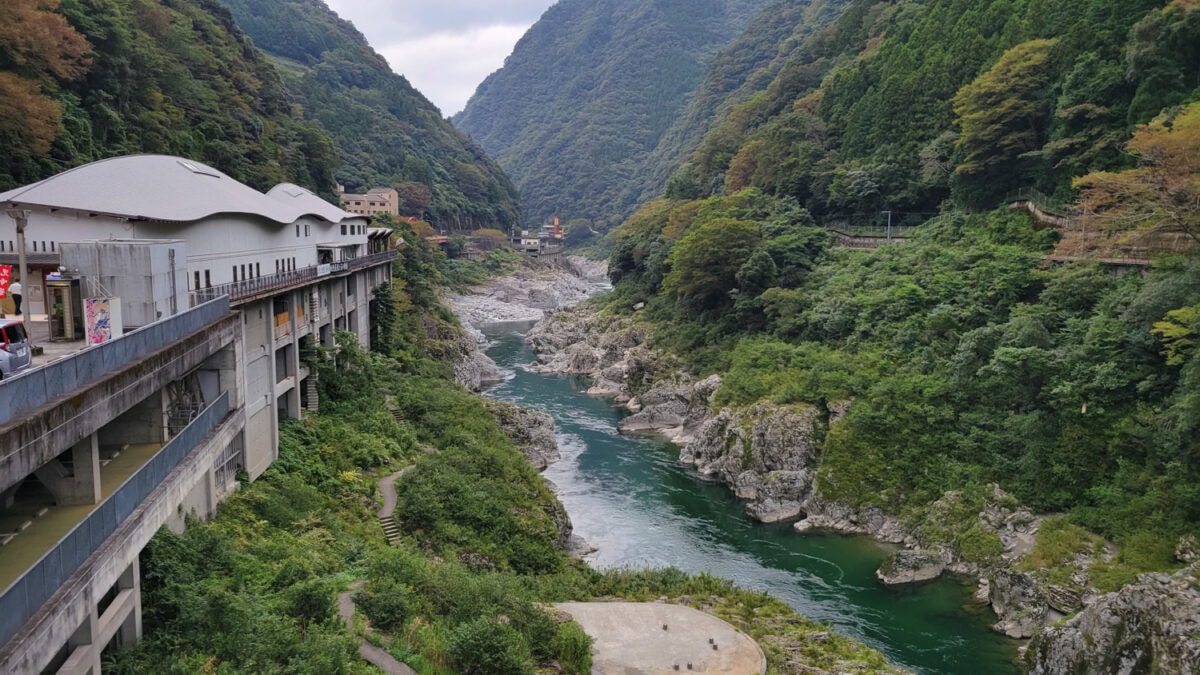The image captures Iya Valley on Shikoku Island, a scenic river valley flanked by steep, forested mountains, with clear green water winding through rocky terrain. On the left, a modern building structure with curved rooftops sits elevated along the hillside, overlooking the river. The lush greenery and rugged landscape create a harmonious blend of natural beauty and subtle human architecture.